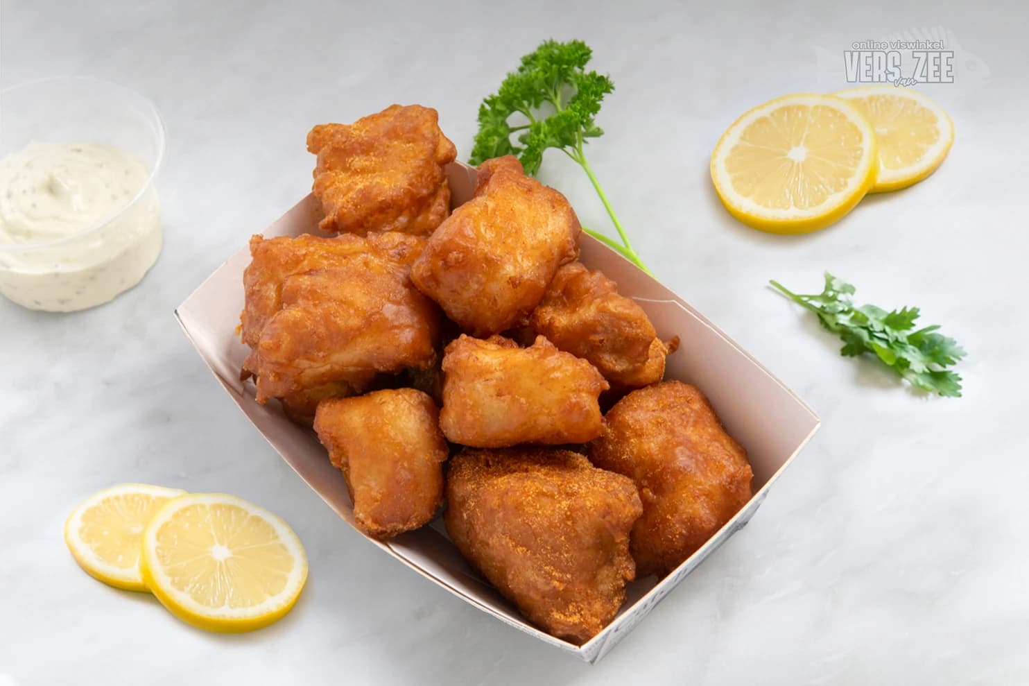 Portie kibbeling (gebakken stukjes vis in beslag) in een bakje, geserveerd met remouladesaus, citroen en peterselie op een lichte marmeren ondergrond.
