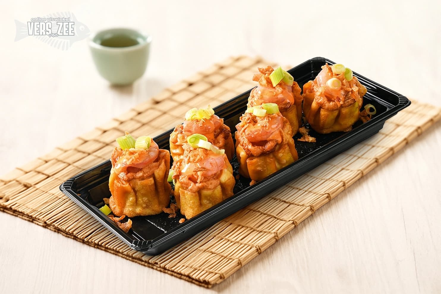 Vijf gefrituurde open dumplings met zalm en garnituur van bosui in zwarte tray, geserveerd op een bamboemat met groene theekop op lichte ondergrond.