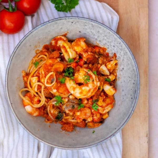 Spaghetti met zeevruchten, waaronder mosselen, venusschelpen en garnalen in tomatensaus, gegarneerd met peterselie in een grijze kom op een gestreepte doek.